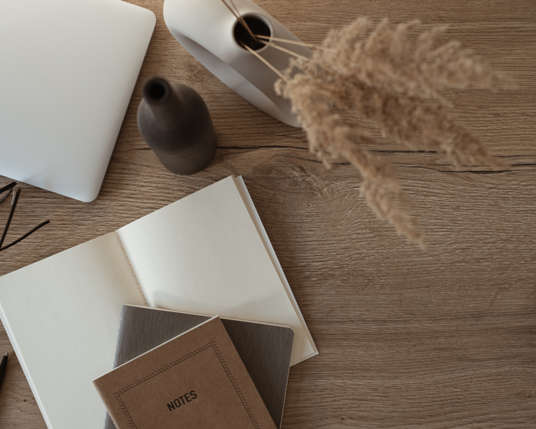 A flatlay of a laptop, ceramic vase, dried pampas grass, and open notebooks on a warm wood desk